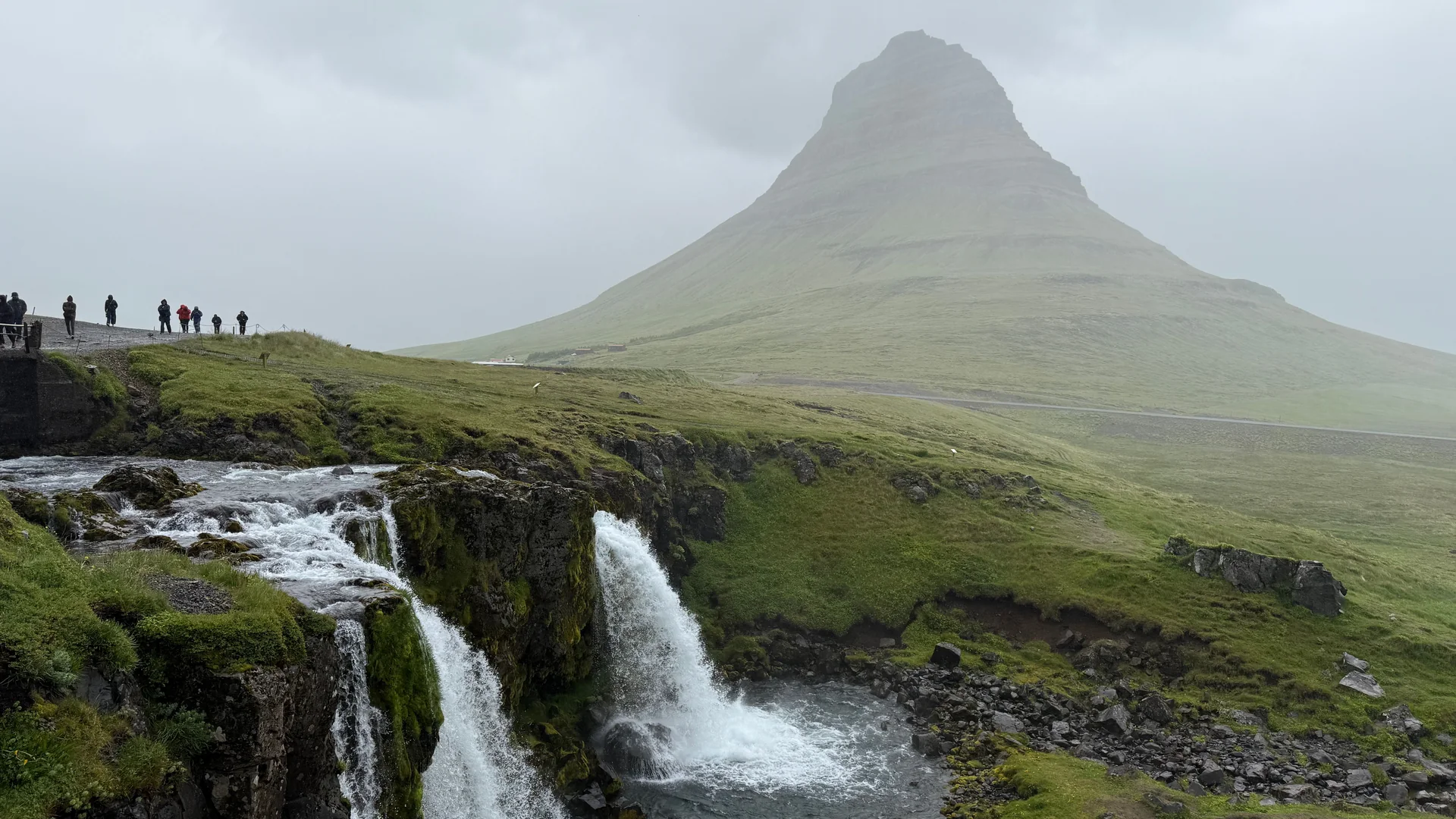 Der Kirkjufell-Berg, der hinter dem Kirkjufellsfoss-Wasserfall auf der Snaefellsnes-Halbinsel aufragt, während Besucher von der Klippenkante aus zuschauen